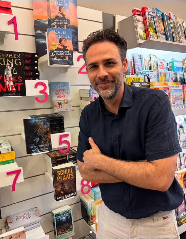Een middeljarige man staat trots in een boekwinkel, zijn eigen boek staat in de kast, op nr 5 van een reeks uitgelichte boeken. 